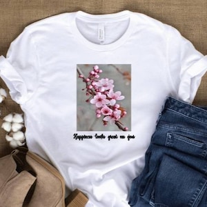 May include: White t-shirt with a floral graphic of pink blossoms on a branch. The text "Happiness looks great on you" is below the graphic. The shirt is paired with blue jeans and beige ankle boots.