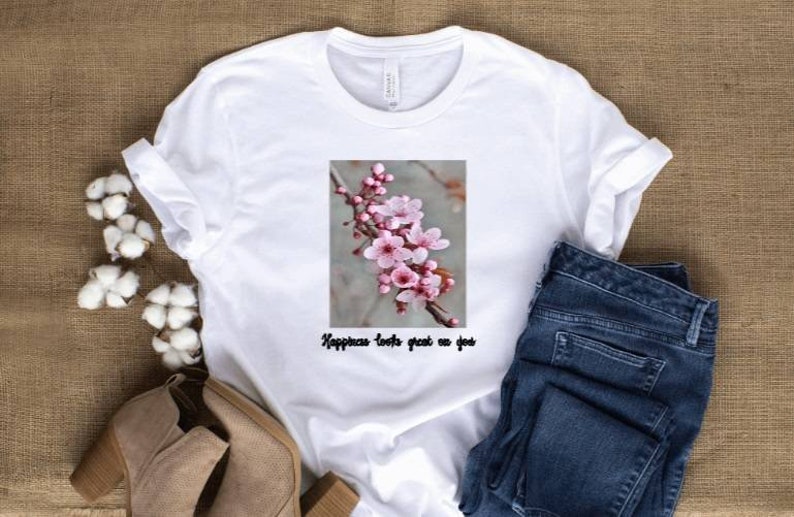May include: White t-shirt with a floral graphic of pink blossoms on a branch. The text "Happiness looks great on you" is below the graphic. The shirt is paired with blue jeans and beige ankle boots.