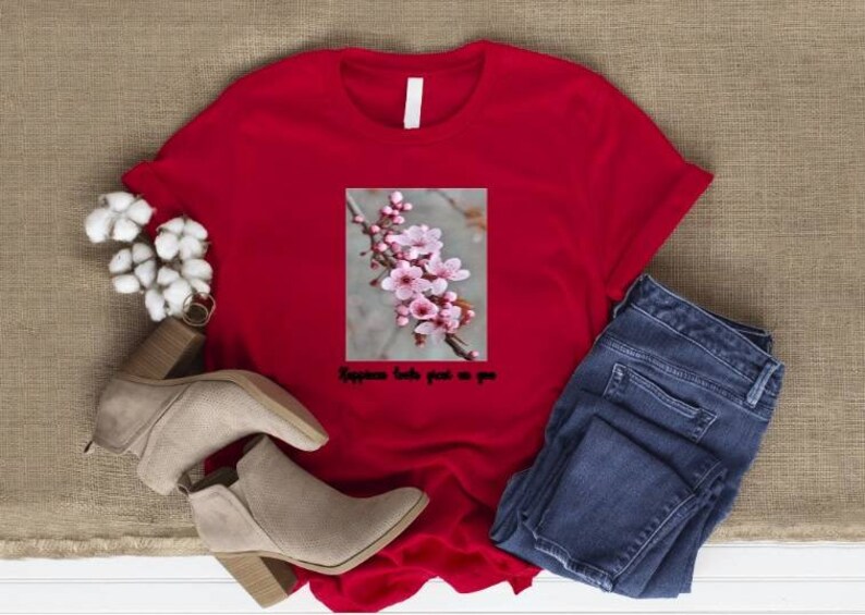 May include: A red t-shirt with a floral graphic and the text "Happiness looks great on you." The shirt is paired with blue jeans and beige ankle boots. Cotton flowers are also in the image.