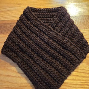May include: A dark brown, hand-crocheted cowl scarf. The scarf is made with a ribbed stitch pattern, creating a textured appearance. The cowl is designed to be worn around the neck and is likely made of a soft, warm yarn.