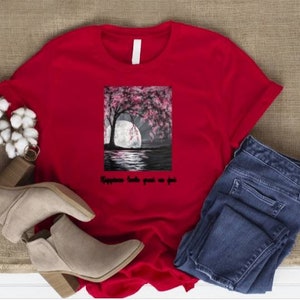 May include: A red t-shirt with a graphic of a tree with pink blossoms, a full moon, and water. The text "Happiness looks good on you" is below the graphic. The shirt is paired with blue jeans and beige ankle boots.