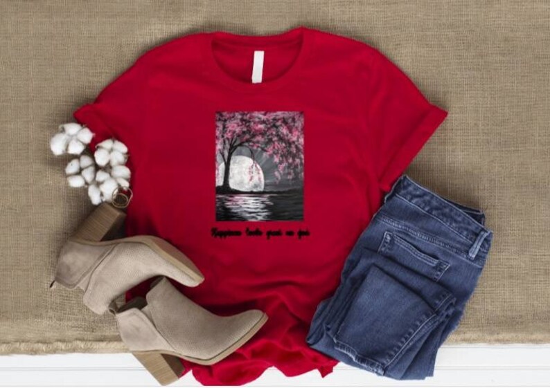 May include: A red t-shirt with a graphic of a tree with pink blossoms, a full moon, and water. The text "Happiness looks good on you" is below the graphic. The shirt is paired with blue jeans and beige ankle boots.