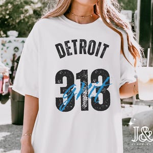 May include: White t-shirt with the text "DETROIT 313 Grit" in black and blue. The shirt has a relaxed fit and short sleeves. The image also includes the J&J Clothing Co. logo.