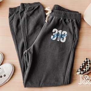 May include: Charcoal gray sweatpants with a drawstring waist and the number "313" printed in white and blue. The sweatpants are laid out on a light wood surface, alongside white sneakers, a baseball cap, and accessories.
