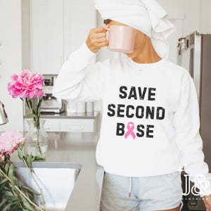 May include: White sweatshirt with the text "SAVE SECOND BASE" in black, with a pink ribbon graphic. The sweatshirt is worn by a person holding a pink mug. Pink peonies are in a vase on a kitchen counter.