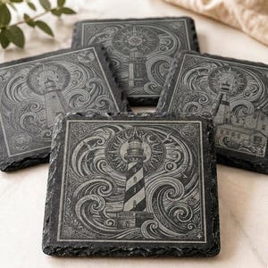May include: A set of five square slate coasters. Each coaster has an etched design of a lighthouse with a sunburst, waves, and birds. The coasters are dark grey with natural, rough edges.