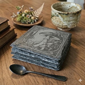 May include: A stack of four square slate coasters with an engraved mushroom design. The coasters are gray and have a rough, natural texture. A metal spoon and a ceramic cup are also visible on the wooden surface.