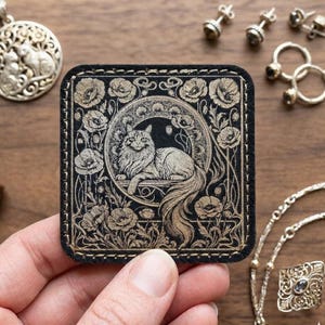 May include: A black and gold square leather patch with a detailed illustration of a cat reclining within a floral design. The patch has a gold border and is stitched with black thread. The image is of a cat-themed accessory.