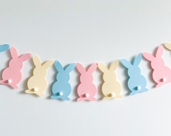 Pode incluir: Uma guirlanda de coelhos da Páscoa em tons pastel. A guirlanda apresenta silhuetas de coelhos em azul claro, rosa e amarelo, cada um com uma cauda de pompom branco. Os coelhos são enfiados em uma corda branca, ideal para decoração de Páscoa.