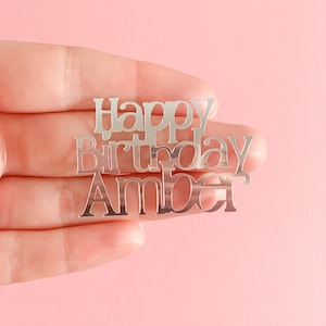 May include: A silver-colored cake topper featuring the text Happy Birthday Amber in a stylized, connected font. The metallic surface is reflective and smooth, with the letters joined together to form a single, cohesive decorative piece.