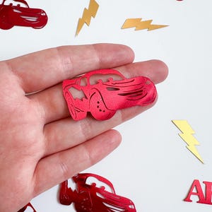 May include: Red car-shaped confetti and gold lightning bolt confetti scattered on a white surface. The name "ARLO" is spelled out in red letters. The confetti is likely for a party or celebration.