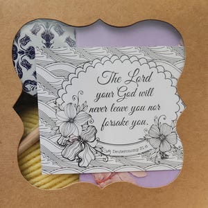 May include: A cardboard gift box with a decorative cutout window revealing a card with the text The Lord your God will never leave you nor forsake you. Deuteronomy 31:6, alongside a ball of yellow yarn and a blue and white patterned item.