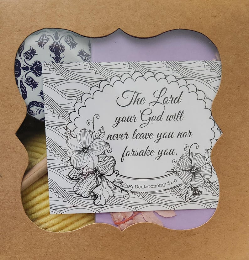 May include: A cardboard gift box with a decorative cutout window revealing a card with the text The Lord your God will never leave you nor forsake you. Deuteronomy 31:6, alongside a ball of yellow yarn and a blue and white patterned item.