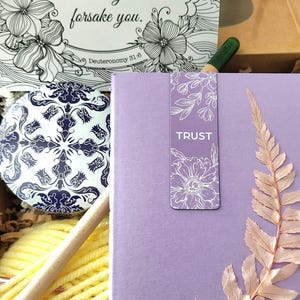 May include: A gift box containing a card with the text The Lord your God will never leave you nor forsake you Deuteronomy 31:6, a blue and white patterned circular tin, a ball of yellow yarn, a wooden crochet hook, and a lavender book with a fern.