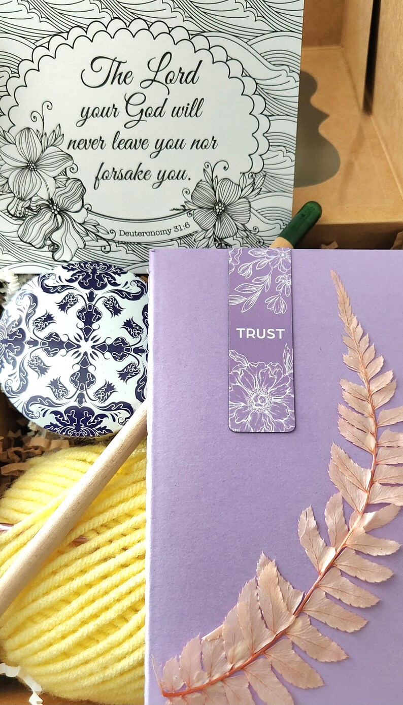 May include: A gift box containing a card with the text The Lord your God will never leave you nor forsake you Deuteronomy 31:6, a blue and white patterned circular tin, a ball of yellow yarn, a wooden crochet hook, and a lavender book with a fern.