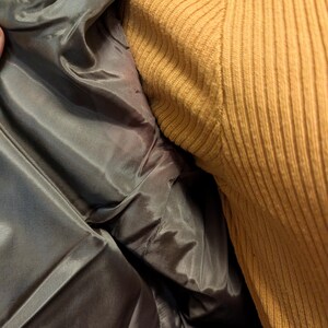 Pu&ograve; includere: Primo piano dell'interno di una giacca grigia, che rivela una fodera liscia e lucida. La giacca &egrave; aperta, mostrando la costruzione interna. Un maglione a coste color senape &egrave; visibile a destra.