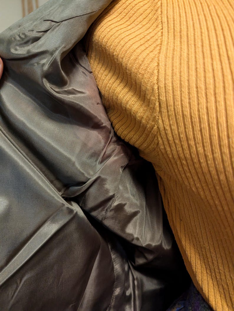 Pu&ograve; includere: Primo piano dell'interno di una giacca grigia, che rivela una fodera liscia e lucida. La giacca &egrave; aperta, mostrando la costruzione interna. Un maglione a coste color senape &egrave; visibile a destra.