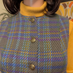 Pu&ograve; includere: Un gilet a quadri grigio, blu, viola e giallo con bottoni color oro. Il gilet &egrave; abbottonato e indossato sopra un dolcevita giallo. Il gilet ha un aspetto classico e su misura.