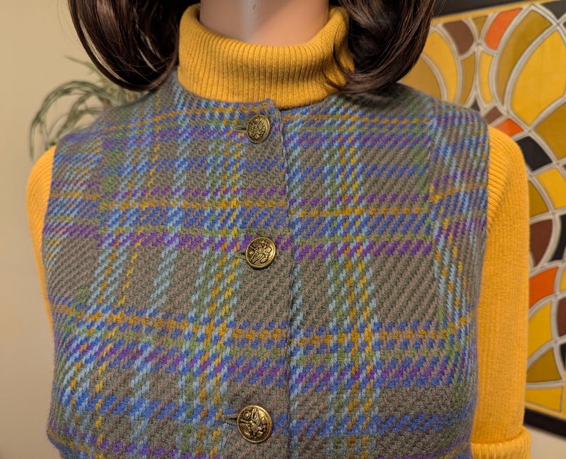 Pu&ograve; includere: Un gilet a quadri grigio, blu, viola e giallo con bottoni color oro. Il gilet &egrave; abbottonato e indossato sopra un dolcevita giallo. Il gilet ha un aspetto classico e su misura.