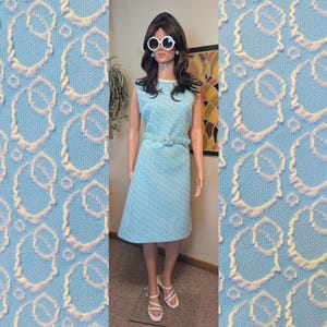May include: A sleeveless, light blue dress with a matching belt. The dress features a repeating pattern and is displayed on a mannequin. The dress is knee-length and has a scoop neckline. The mannequin is wearing white sunglasses and white sandals.