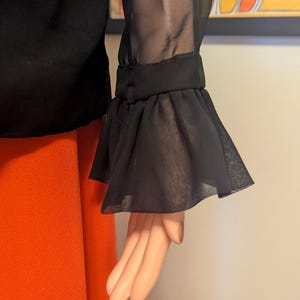 May include: Black blouse with sheer sleeves and ruffled cuffs. The blouse is paired with orange pants. The sleeve detail is the focal point, with the sheer fabric and ruffled edge adding a touch of elegance to the garment.