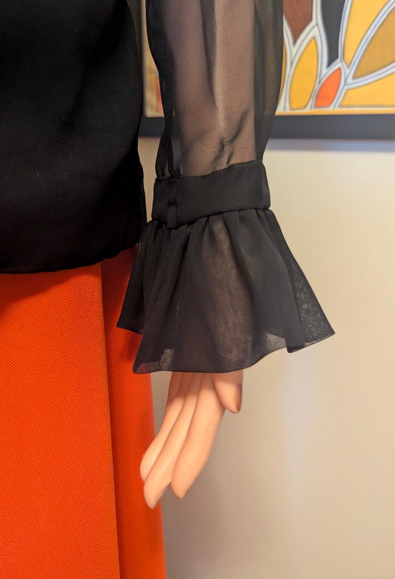 May include: Black blouse with sheer sleeves and ruffled cuffs. The blouse is paired with orange pants. The sleeve detail is the focal point, with the sheer fabric and ruffled edge adding a touch of elegance to the garment.