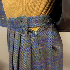 Pu&ograve; includere: Primo piano di una gonna in tessuto a quadri grigio, blu, viola e giallo. La gonna ha una chiusura con cerniera e bottone. La parte superiore &egrave; una maglia a coste giallo senape.
