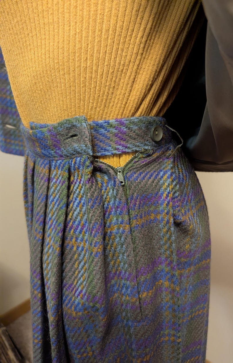Pu&ograve; includere: Primo piano di una gonna in tessuto a quadri grigio, blu, viola e giallo. La gonna ha una chiusura con cerniera e bottone. La parte superiore &egrave; una maglia a coste giallo senape.