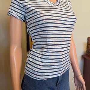 May include: A white striped short-sleeved shirt with blue, black, and red stripes. The shirt has a v-neck and a button closure at the neckline.