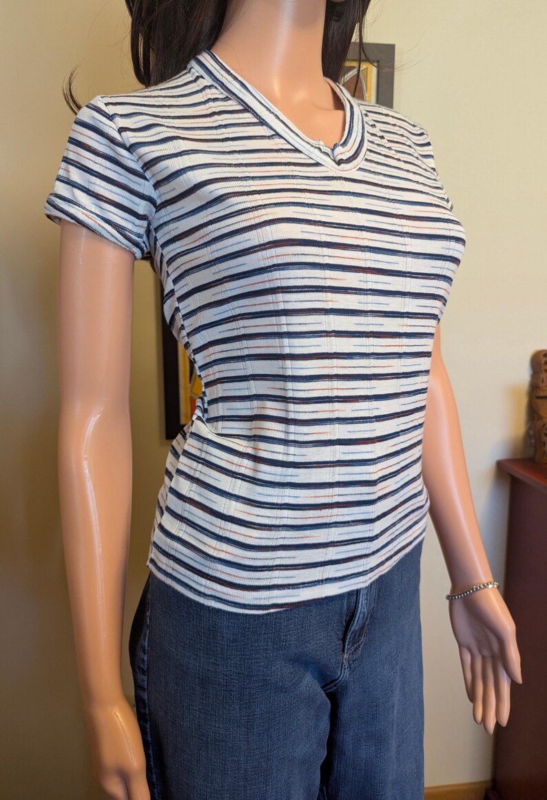 May include: A white striped short-sleeved shirt with blue, black, and red stripes. The shirt has a v-neck and a button closure at the neckline.