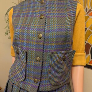 Pu&ograve; includere: Un completo vintage composto da gilet e gonna in tweed con motivo a quadri grigio, blu, viola e giallo. Il gilet ha quattro bottoni dorati decorativi e due tasche frontali. L'outfit &egrave; abbinato a un dolcevita giallo.
