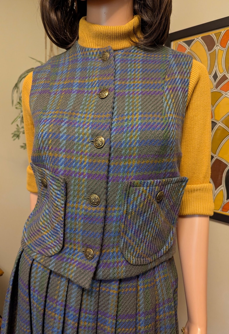 Pu&ograve; includere: Un completo vintage composto da gilet e gonna in tweed con motivo a quadri grigio, blu, viola e giallo. Il gilet ha quattro bottoni dorati decorativi e due tasche frontali. L'outfit &egrave; abbinato a un dolcevita giallo.