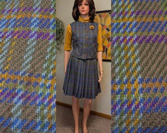 May include: A vintage three-piece outfit on a mannequin. The outfit includes a pleated skirt, a vest with pockets, and a long-sleeved mustard yellow top. The vest and skirt are made of a plaid fabric with blue, green, purple, and yellow hues. A gold-colored brooch adorns the vest.