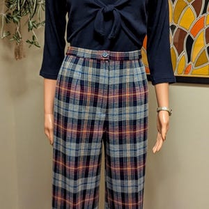 Op de afbeelding: Een mannequin draagt een donkerblauw lang mouw shirt met een strik aan de voorkant en een geruite broek. De broek heeft een blauw, rood en wit geruit patroon.