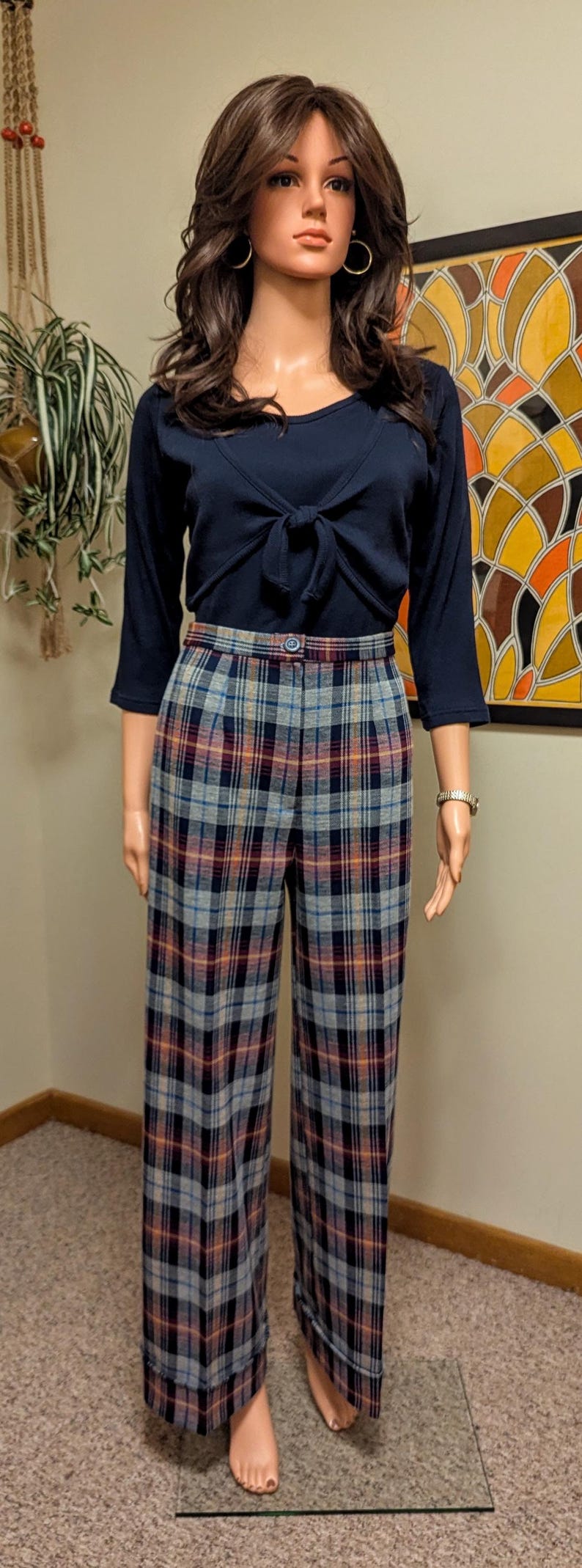 Op de afbeelding: Een mannequin draagt een donkerblauw lang mouw shirt met een strik aan de voorkant en een geruite broek. De broek heeft een blauw, rood en wit geruit patroon.