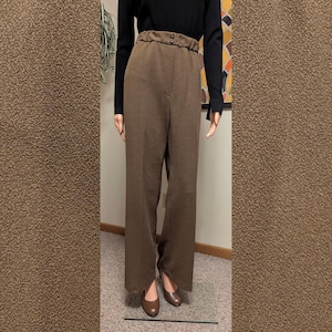 May include: Brown high-waisted trousers with a wide leg. The pants have a button closure and a tailored fit. The model is wearing a black long-sleeve top and brown heels. The pants are made of a textured fabric.