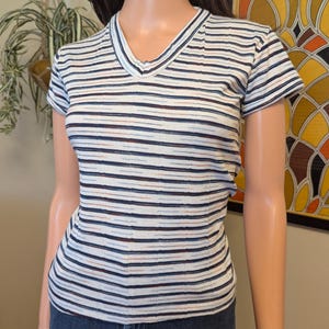 May include: White striped short-sleeved shirt with thin blue, brown, and red stripes. The shirt has a v-neck.