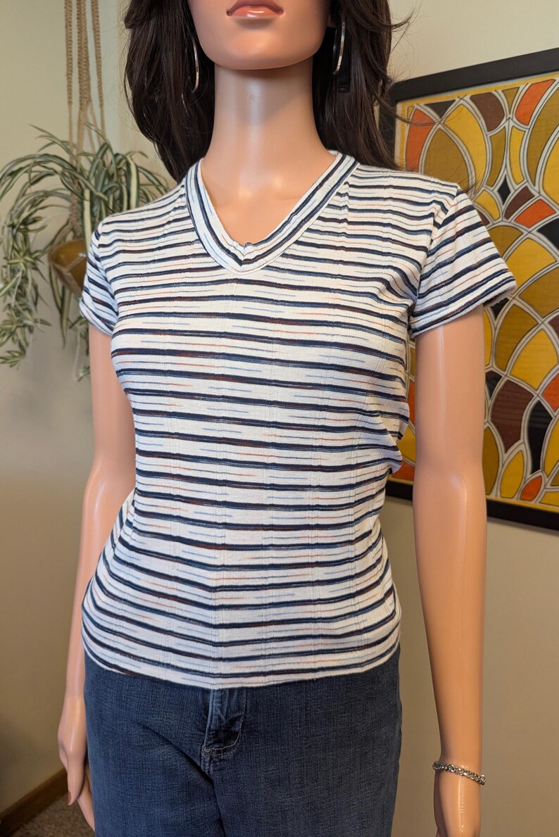 May include: White striped short-sleeved shirt with thin blue, brown, and red stripes. The shirt has a v-neck.