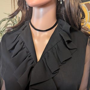May include: Black chiffon blouse with a ruffled collar and sheer sleeves. A black choker necklace is worn around the neck. The blouse has a V-neckline and is displayed on a mannequin.