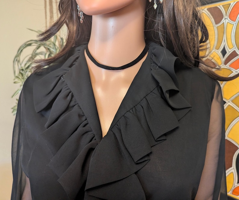 May include: Black chiffon blouse with a ruffled collar and sheer sleeves. A black choker necklace is worn around the neck. The blouse has a V-neckline and is displayed on a mannequin.