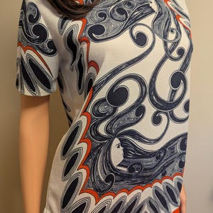Pu&ograve; includere: Top bianco a maniche corte con un motivo ripetuto di volti femminili stilizzati e capelli fluenti in blu navy e rosso. Il top &egrave; abbinato a una gonna arancione, creando un contrasto sorprendente.
