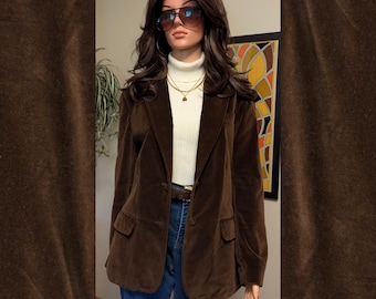 May include: A dark brown velvet blazer with a notched lapel, two front buttons, and two flap pockets. The blazer is worn over a white ribbed turtleneck sweater, a gold chain necklace, and blue denim jeans, paired with gradient sunglasses.