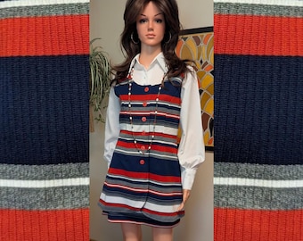 May include: A sleeveless mini dress featuring horizontal stripes in navy blue, red, white, and gray. The dress has a button-down front with red buttons and is worn over a long-sleeved white collared shirt. A long beaded necklace hangs over the dress.