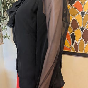 May include: Black sheer blouse with ruffled collar and cuffs. The long sleeves are made of a sheer material. The blouse is paired with a bright red skirt. The blouse has a button-down front.