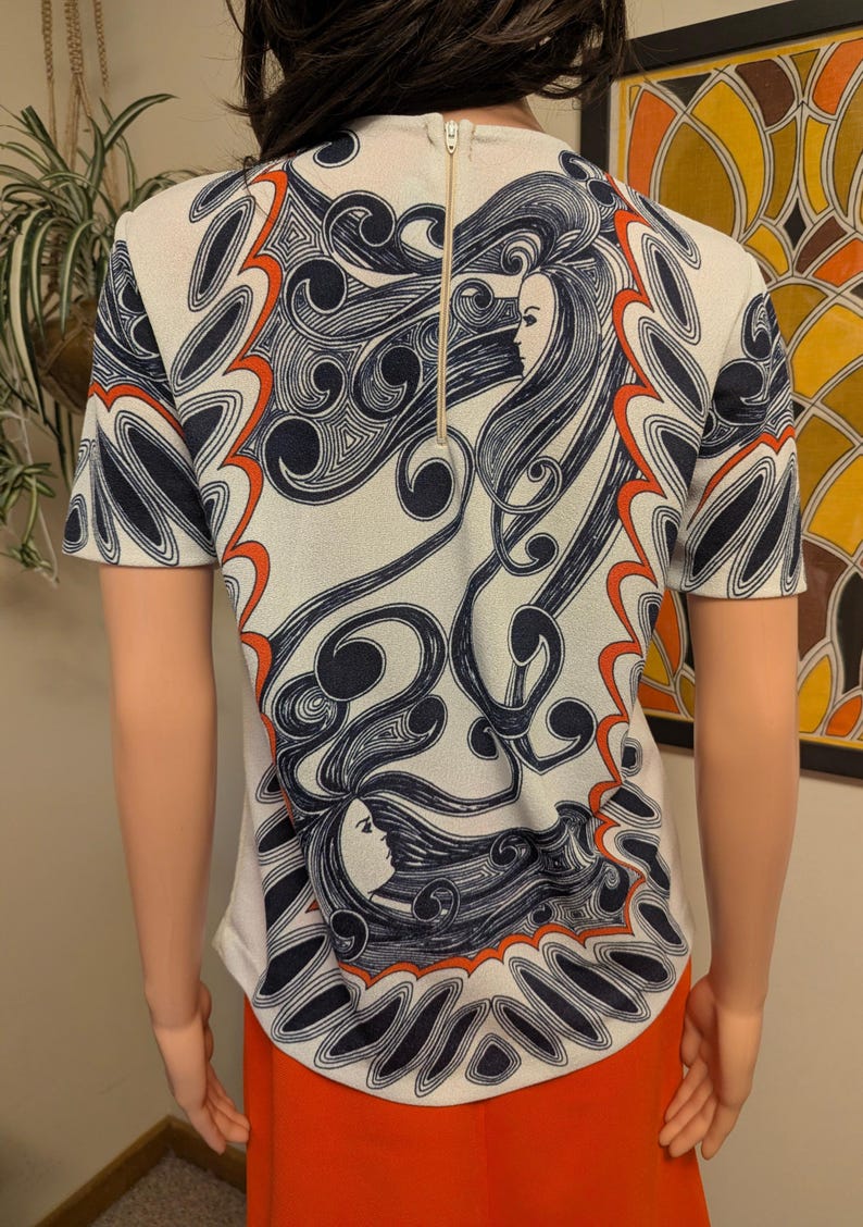 Pu&ograve; includere: Top color crema a maniche corte con un design sorprendente con motivi a spirale blu navy e arancioni e il profilo di un volto femminile. Il top ha una cerniera dorata sul retro.