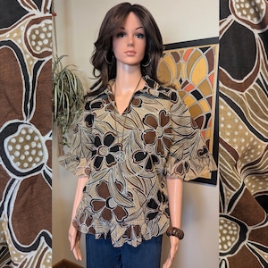 May include: A floral print blouse with a V-neck and ruffled sleeves. The blouse features a brown, black, and beige floral pattern. The mannequin is wearing blue jeans and a necklace with a peace sign pendant. The blouse is a vintage style.