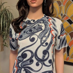 Pu&ograve; includere: Un top vintage a maniche corte con sfondo bianco e un motivo ripetuto di volti femminili stilizzati e capelli fluenti in blu navy e arancione. Il top &egrave; abbinato a una gonna arancione.