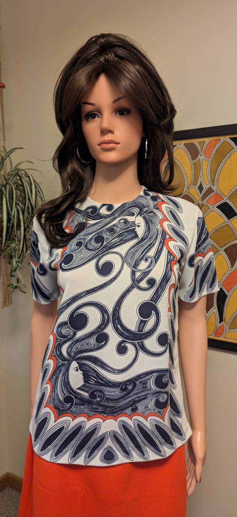 Pu&ograve; includere: Un top vintage a maniche corte con sfondo bianco e un motivo ripetuto di volti femminili stilizzati e capelli fluenti in blu navy e arancione. Il top &egrave; abbinato a una gonna arancione.
