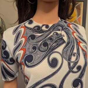 Pu&ograve; includere: Top bianco a maniche corte con un design sorprendente. Il motivo presenta linee fluenti in blu navy e arancione, con un'immagine centrale del profilo di una donna. Il top ha uno scollo tondo e una silhouette aderente, che mostra un'estetica vintage.