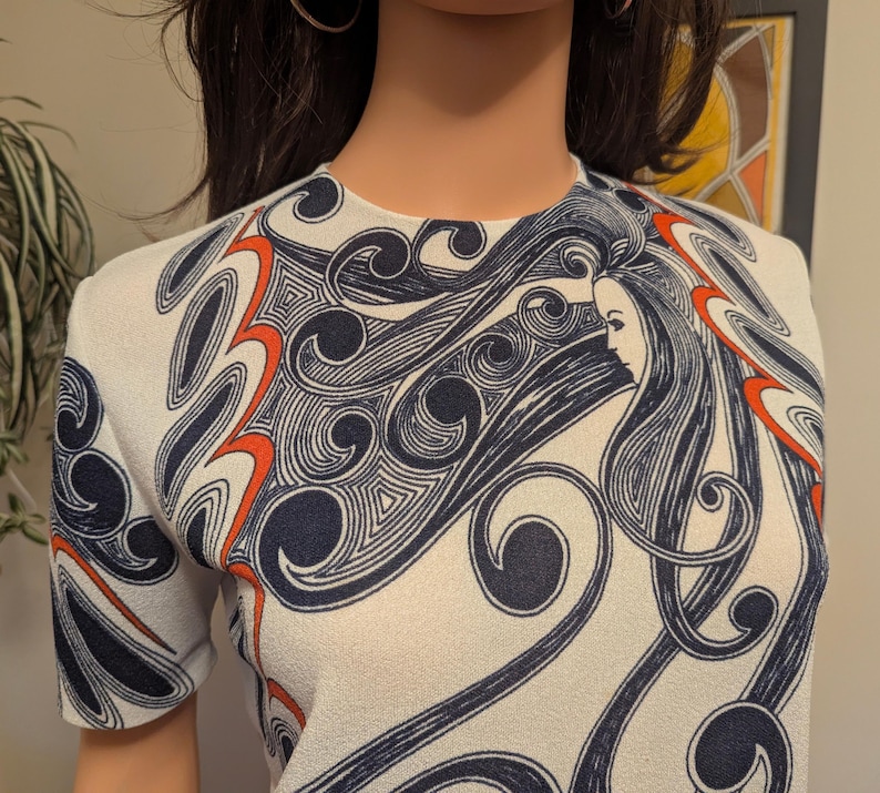 Pu&ograve; includere: Top bianco a maniche corte con un design sorprendente. Il motivo presenta linee fluenti in blu navy e arancione, con un'immagine centrale del profilo di una donna. Il top ha uno scollo tondo e una silhouette aderente, che mostra un'estetica vintage.